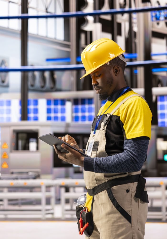 Male engineer analyzing industry 4.0 system performance in smart manufacturing plant. Black man monitoring machinery and robotic arms, ensuring optimal production and industrial automation efficiency.